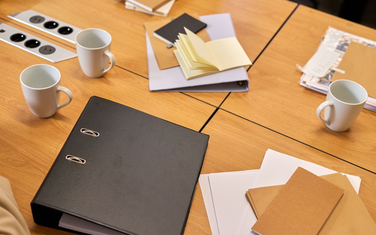 A binder, loose papers and two mugs on a wooden meeting table