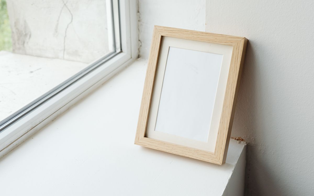 An empty light wood picture frame leaning against a wall on a windowsill