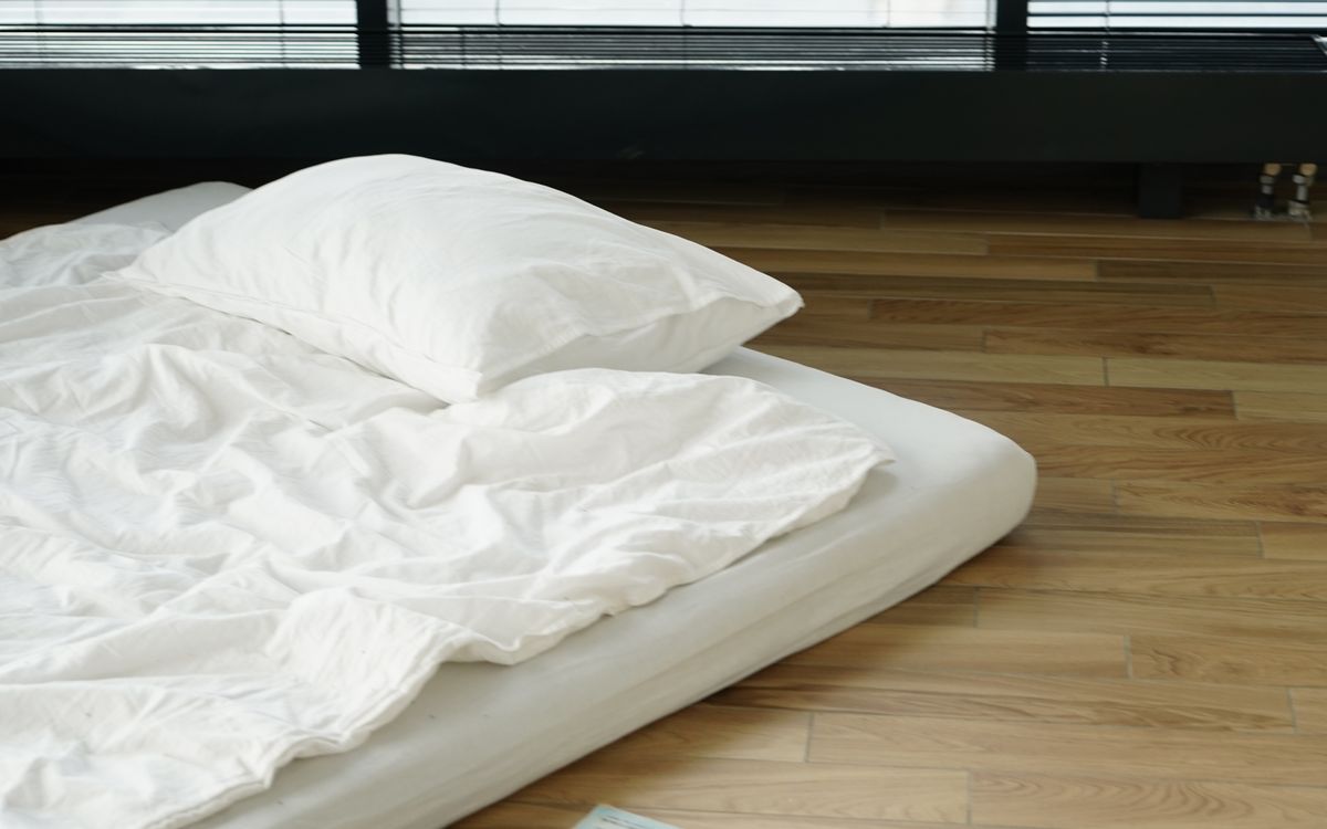 A mattress made up with white bedding on a bare wooden floor by a window
