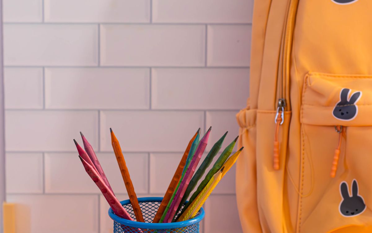A cup of colored pencils beside a yellow backpack against a white tiled wall